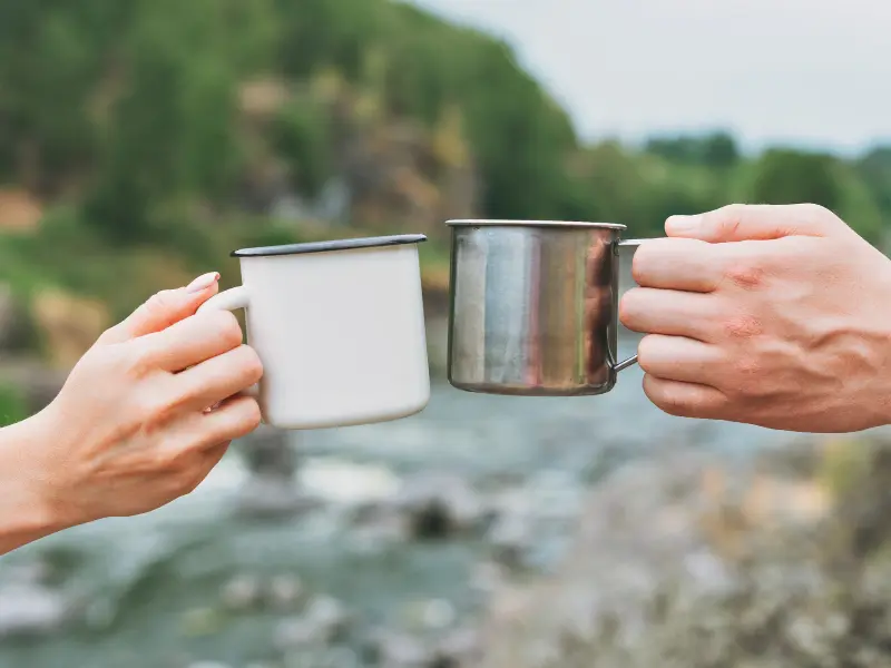 Two enamel mugs clinking at a riverside campsite.