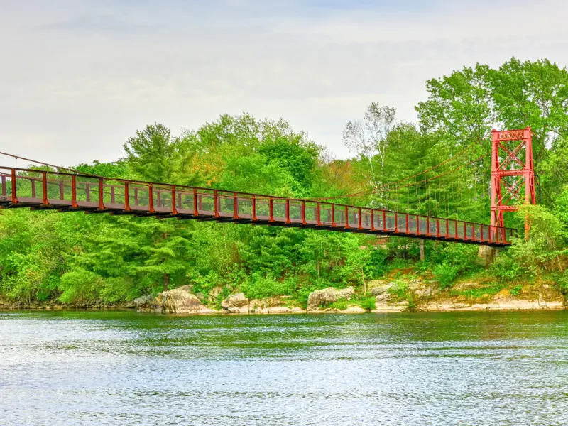 Red suspension footbridge over the Androscoggin River surrounded by trees.