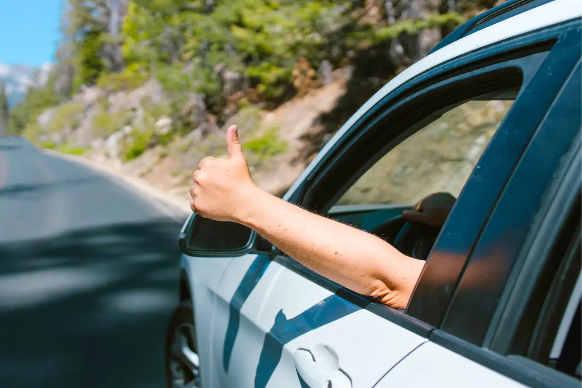 Person giving a thumbs-up out of a car window on a forested road.