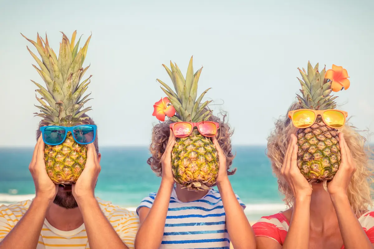 Three people holding pineapples with sunglasses in front of the ocean, thinking of fun places to go in the summer.
