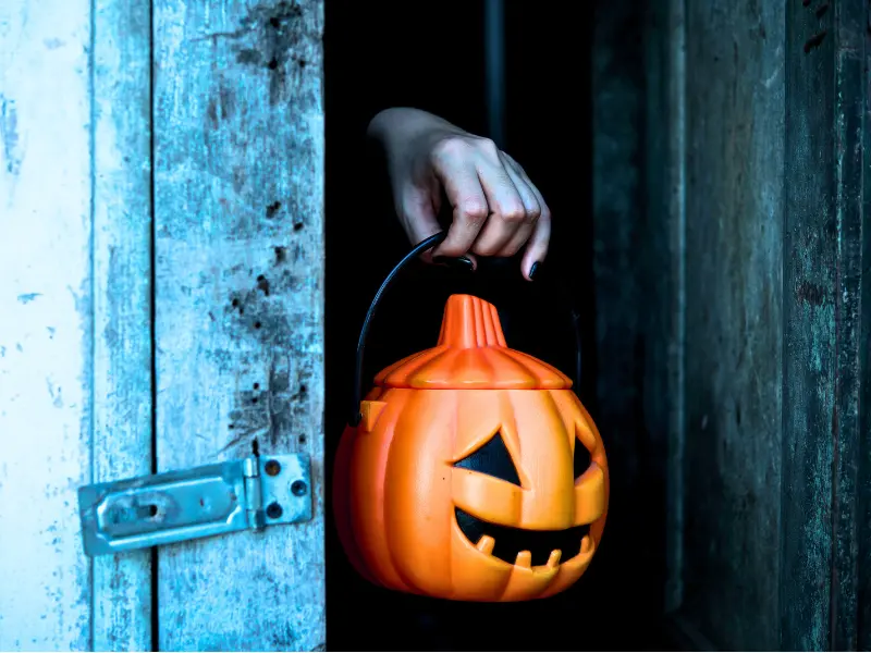 Hand holding a plastic pumpkin bucket in a spooky doorway.