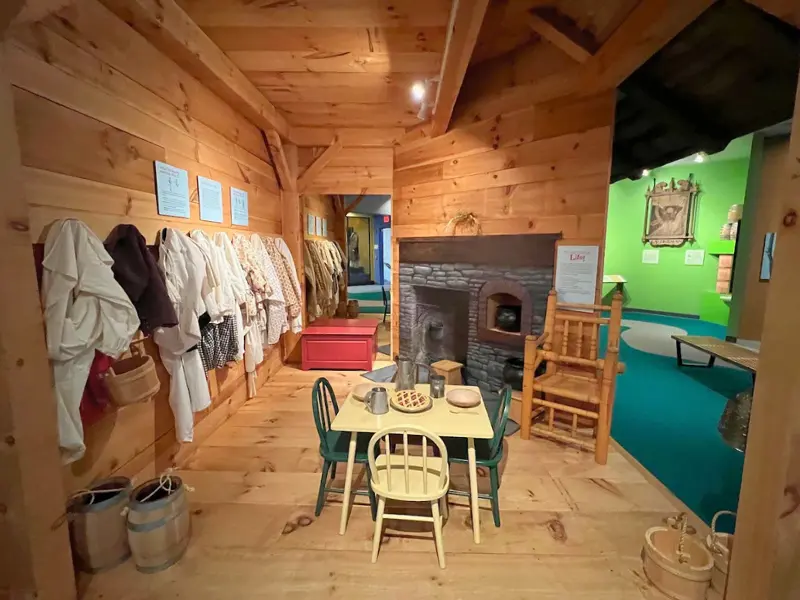 Kids colonial dress-up area with wooden walls, costumes, and a table set inside the museum.