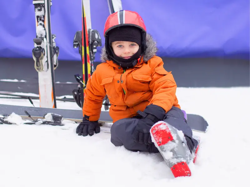 Young child sitting in snow wearing ski boots and helmet at ski resort